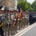 Line of soldiers in uniform standing at attention during a parade, one holding a blue-yellow-red flag.
