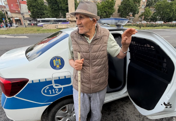 Older man with a cane and hat stands by the open rear door of a white and blue police car on a city street.