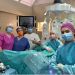 Surgical team in masks and gowns pose in an operating room beside a robotic surgery system with multiple robotic arms.