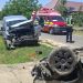 Two damaged vehicles after a crash: a silver van with severe front-end damage on the grass and a flipped car on the road, with paramedics and ambulances nearby on a sunny day.