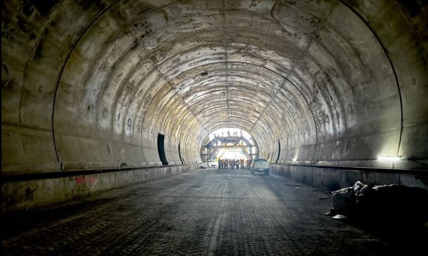 Long concrete tunnel under construction; roadbed graded and workers gathered near the bright far end exit.