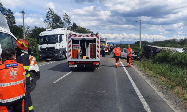 Emergency responders in orange vests at a highway accident scene with a tipped vehicle on the roadside and police/ambulance nearby