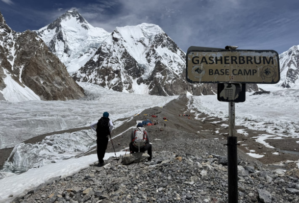 Climbers on a rocky, snow-dusted trail with a Gasherbrum Base Camp sign and towering snowy peaks in the background.