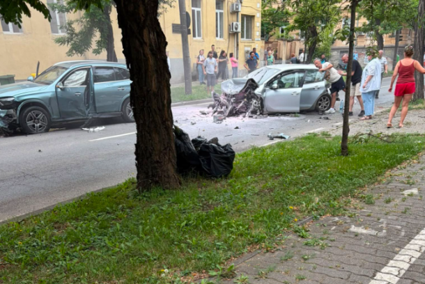 Two cars damaged in a street collision with debris on the road and a crowd of bystanders watching nearby on a residential street.