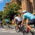 Cyclists ride in a group along a sunny city street past a red-brick historic building with green trees nearby.