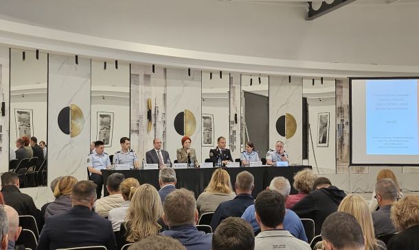 Panel of speakers seated at a long table on a stage, with nameplates and microphones, addressing a seated audience in a conference room.