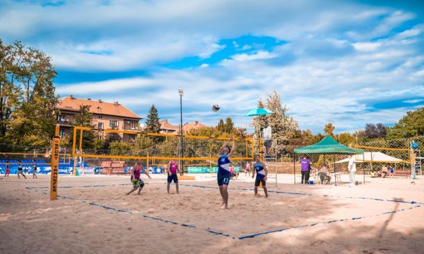 Beach volleyball match on a sunny sandy court with players jumping near the net and a ball in mid-air; spectators and tents in the background.