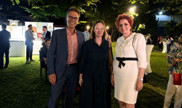 Three adults posing at an outdoor evening reception; man in a blue suit with pink shirt, two women in black and white dresses, string lights and attendees in the background.