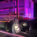 Nighttime crash scene: a black car has collided with the side of a large trailer truck, the driver's door is open and interior is visible with purple emergency lights.