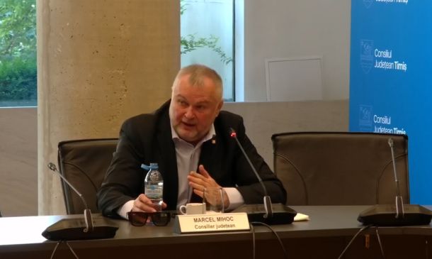 Man in a dark suit speaks into a microphone at a panel, nameplate reads 'Marcel Mihoc' and 'Consilier județean' beside a bottle of water on the table, with a blue council backdrop behind him