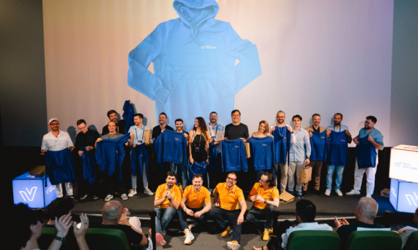 Group of people on a stage posing with blue jackets, in front of a large projected image of a blue hoodie on the screen behind them, an event photo showing staff and speakers holding up jackets.