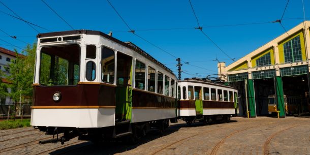 Vintage tram cars parked on cobblestone tracks in a rail yard under a bright blue sky, beside a museum depot with green doors.