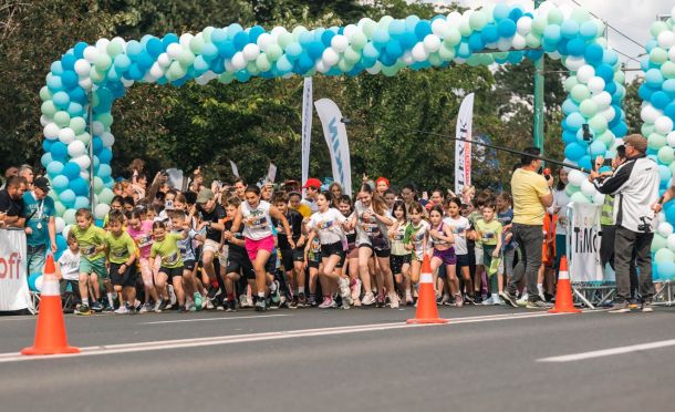 Children start a road race under a blue‑green balloon arch; spectators and organizers watch nearby.
