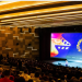 A crowded theater auditorium with attendees looking toward a brightly lit stage and a large colorful screen displaying festival graphics on the right side.