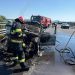 Firefighters in turnout gear extinguish a burned car on a highway, smoke rising, with a red fire engine in the background and a wet, reflective road surface.