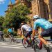 Cyclists ride in a group along a sunny city street past a red-brick historic building with green trees nearby.